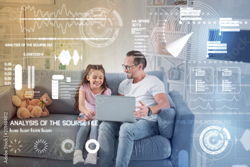 Funny cartoon. Cheerful emotional young father sitting comfortably on the sofa and holding a modern laptop while watching a funny cartoon with his cute daughter