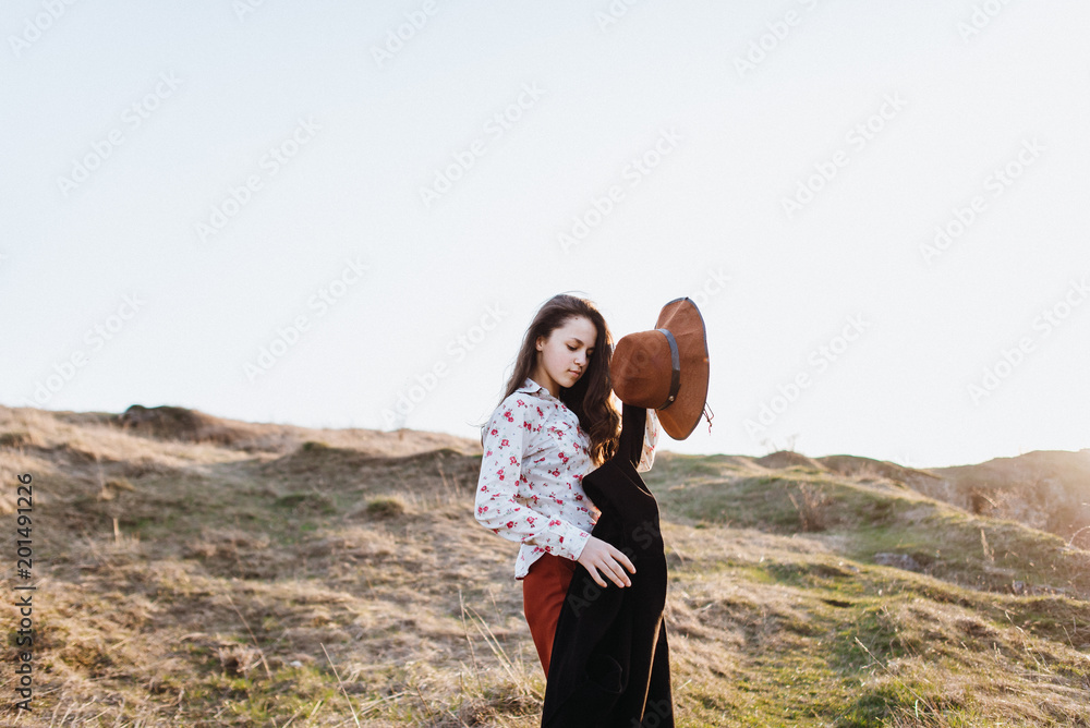 Naklejka premium Beautiful girl in brown pants and with a hat walks to mountain places with a young spring grass. traveler hipster is holding hat in sunset. travel concept