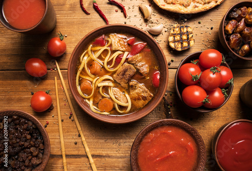 Asian beef soup in wooden rustic background. Traditional Uzbek dish is Lagman. Uzbek flatbread lavash, garlic, tomatoes, noodles, red pepper in frame. Flat lay. Top view.