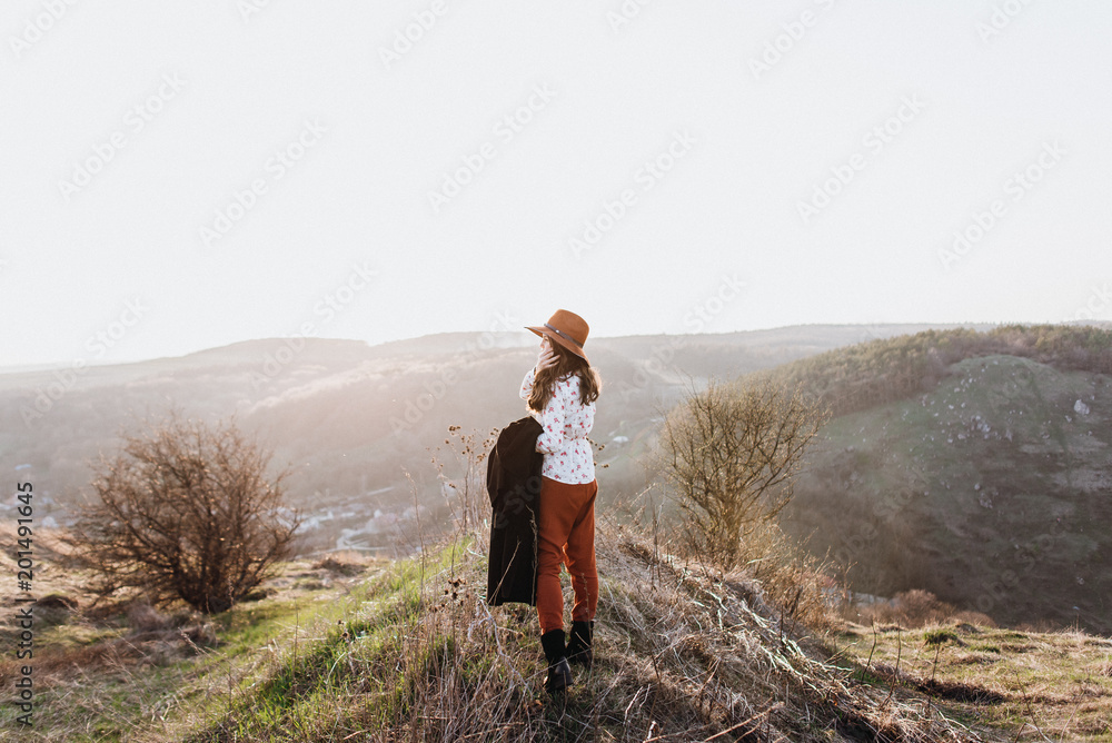Beautiful girl in brown pants and with a hat walks to mountain places with a young spring grass. traveler hipster is holding hat in sunset. travel concept. View from behind