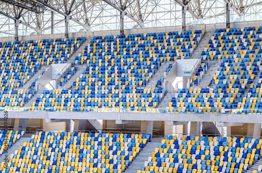 Fototapeta premium Siedzący kibice kolorowe plastikowe krzesła na tle stadionu piłkarskiego. Pusty stadion boisko do piłki nożnej zielona trawa na arenie lekkoatletycznej piłki nożnej.