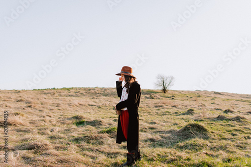 Beautiful girl in a black coat, brown pants and with a hat walks to mountain places with a young spring grass. traveler hipster holding hat in sunset. Wearing stylish fall outfit. close-up portrait
