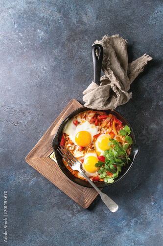 Traditional Israeli Cuisine dishes Shakshuka. Fried egg with vegetables tomatoes and paprika in cast-iron pan on wooden board with cloth and herbs over blue texture background. Top view, space. photo