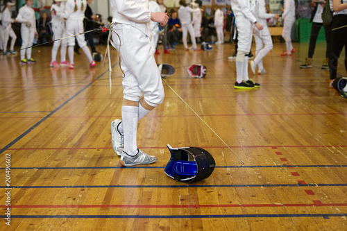 Girl participant in the fencing competition on swords is in the center of gym holding a sword in her hand, waiting for next battle. Fencing mask is on the floor © Slava