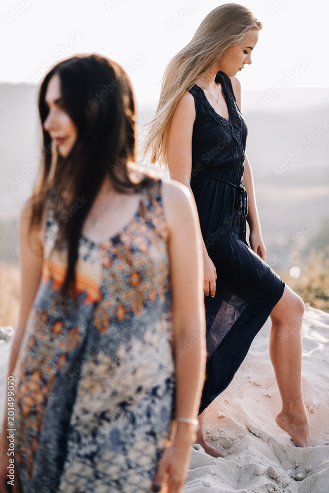 Fototapeta premium beautiful young girls walking in the light of long summer dresses for mountainous terrain, sand and posing. the wind and the sunset