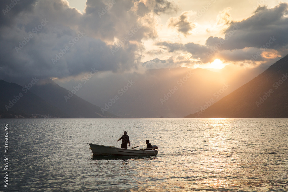 Two people sailing on wooden boat. Men are fishers. Amazing sunset landscape with sea, mountains, colorful orange and blue cloudy sky. Beautiful The Boka Kotor Bay view in Montenegro.