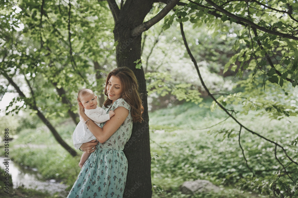 Cute smiling young mother with long hair holding a pretty baby in a white dress. Mother looks tenderly at the child. They are in the green summer forest. Cute smiling young mother with long hair holding a pretty baby in a white dress. Mother looks tenderly at the child. They are in the green summer forest.