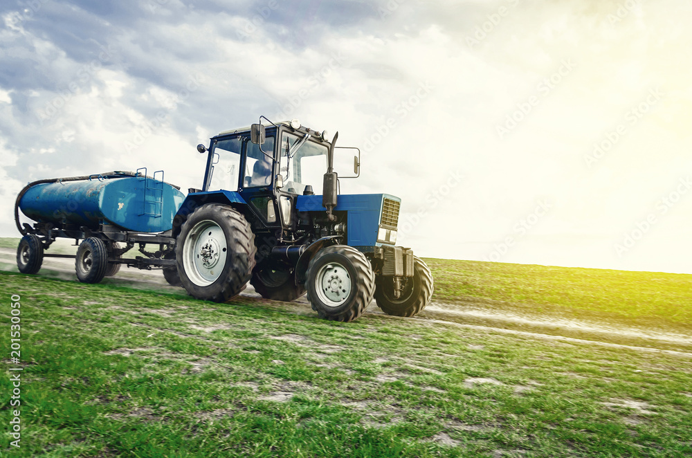 Obraz premium tractor of blue color with a barrel trailer rides along the spring field along the road