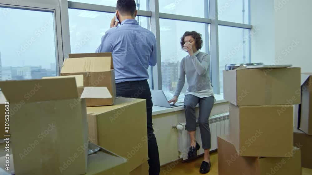 Zoom in young businesswoman sitting on window sill in new office with stacks of cardboard boxes, working on laptop and drinking coffee while her male colleague talking on cell phone Zoom in young businesswoman sitting on window sill in new office with stacks of cardboard boxes, working on laptop and drinking coffee while her male colleague talking on cell phone