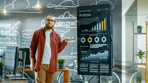 Male Developer Looks at Interactive Whiteboard with Charts, Graphs and Growth Statistics. He Points at Display with Presenting Gesture and Smile. In the Background Creative Office with Chalkboard Wall photo