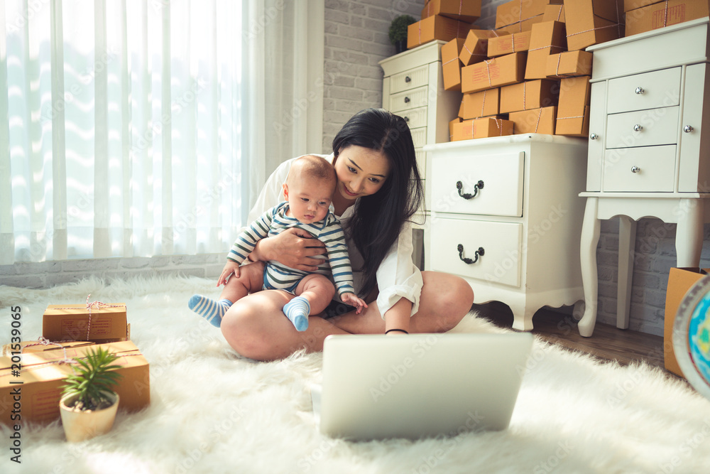 Beautiful woman business mom is taking packaging and laptop with baby boy and work at home concepts. - obrazy, fototapety, plakaty 