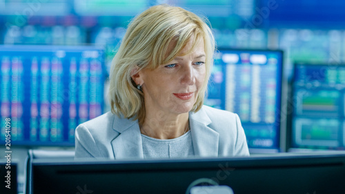 Portrait of the Senior Female Stock Trader Operating at Her Workstation. Behind Her Multiple Monitors Showing Data, Ticker Numbers and Graphs.