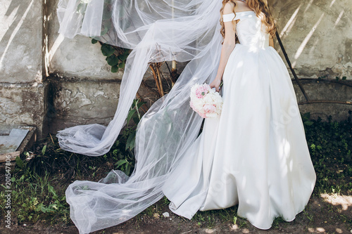 stylish bride with peony bouquet and long veil, posing in sunny garden on wedding day. luxury wedding girl, happy newlywed in silk gown. romantic moments