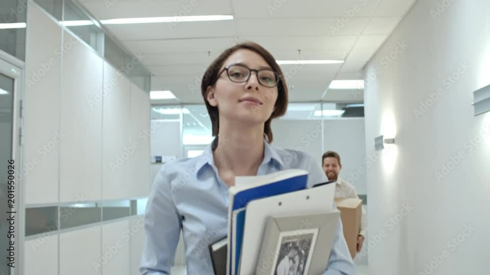 Young attractive businesswoman walking through hallway in new office center with personal belongings and smiling while looking at excited colleagues running with cardboard boxes Young attractive businesswoman walking through hallway in new office center with personal belongings and smiling while looking at excited colleagues running with cardboard boxes