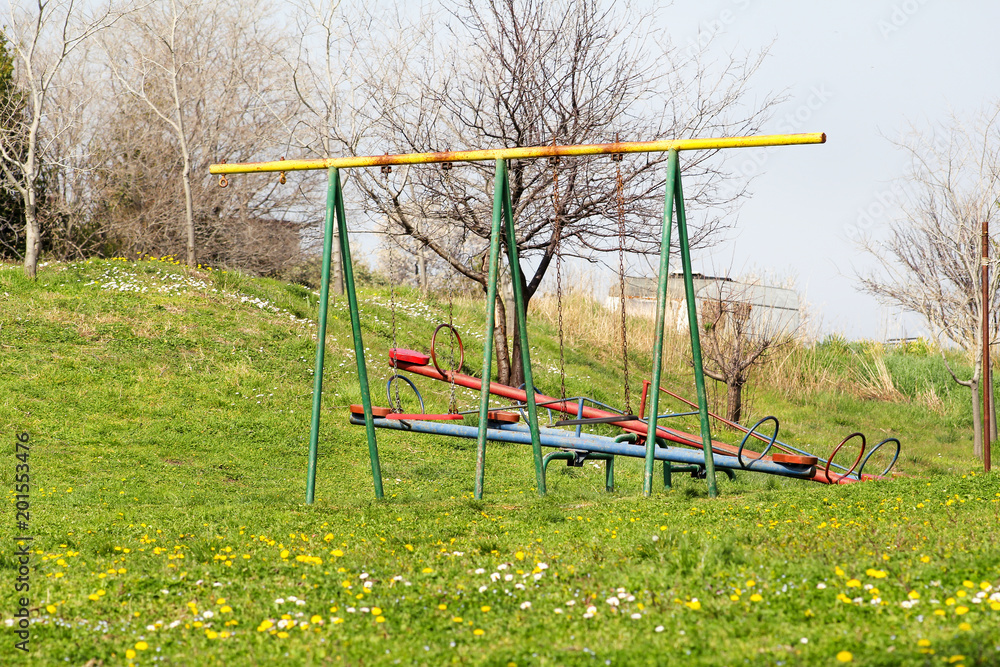 Two empty children swings and seesaw at playground activities in public park / Beautiful natural environment, grass, spring flowers / Colorful playground equipment for children / Sunny day in spring. Two empty children swings and seesaw at playground activities in public park / Beautiful natural environment, grass, spring flowers / Colorful playground equipment for children / Sunny day in spring.
