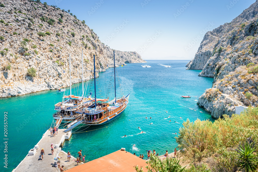 Fototapeta premium Beautiful sunny coast view to the greek blue sea with crystal clear water beach with big tourist boat cruising around pier in Vathys Bay , Patmos Island, Kos, Dodecanese Islands, Greece