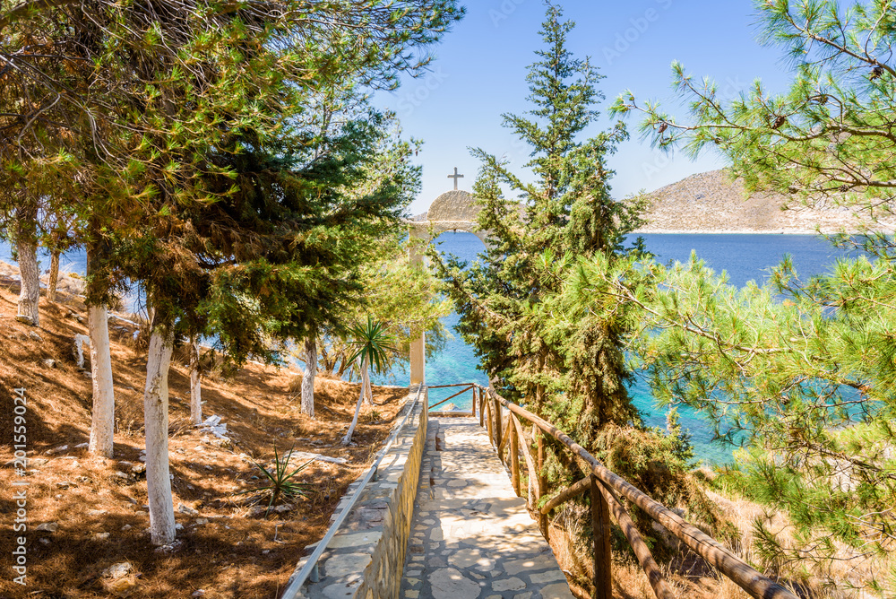 Fototapeta premium Beautiful sunny coast view to the greek blue sea with crystal clear water beach near a small chapel with a white arch with holy cross situated in a small forest, Leros, Dodecanese Islands/ Greece