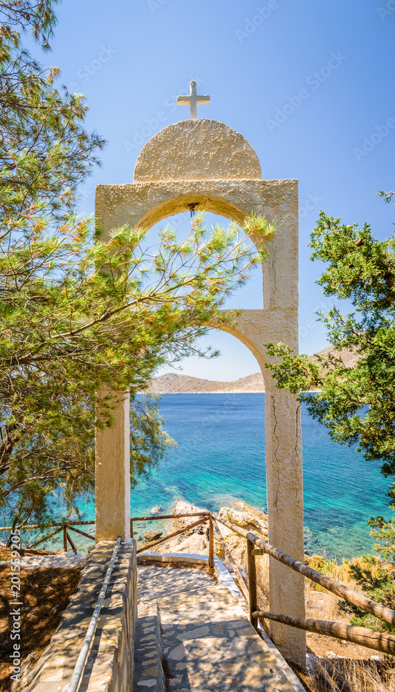 Beautiful sunny coast view to the greek blue sea with crystal clear water beach near a small chapel with a white arch with holy cross situated in a small forest, Leros, Dodecanese Islands/ Greece