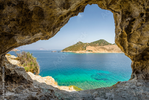 Fototapeta Naklejka Na Ścianę i Meble -  Beautiful sunny view through a rocky cave to the greek blue sea with crystal clear water from an a hill with boats cruising fishing surrounded by mountains, Patmos Island, Kos, Dodecanese/ Greece 