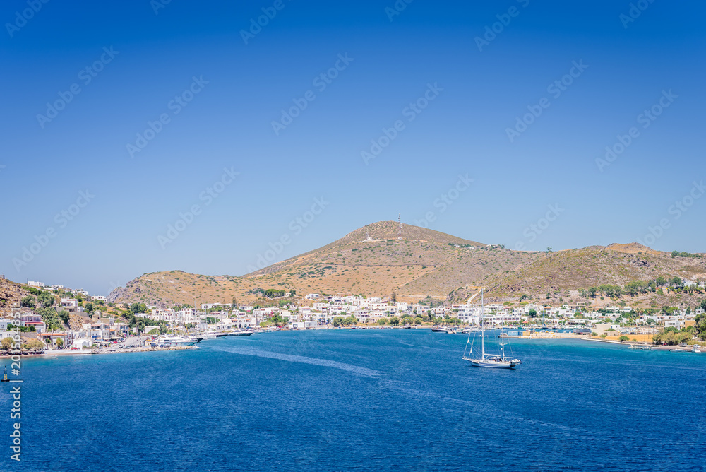Skala, Patmos Island, Dodecanese, Greece: Beautiful sunny sunset greek village town harbor view white church to the aegean sea with crystal clear water surrounded by mountains and ferry in background