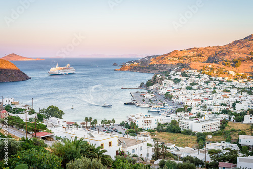 Fototapeta Naklejka Na Ścianę i Meble -  Skala, Patmos Island, Dodecanese, Greece: Beautiful sunny sunset greek village town harbor view white church to the aegean sea with crystal clear water surrounded by mountains and ferry in background