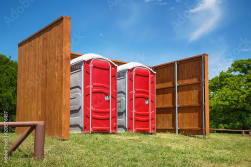 Two red portable restrooms with a wood fence around them in a park. Trees and blue sky background.
