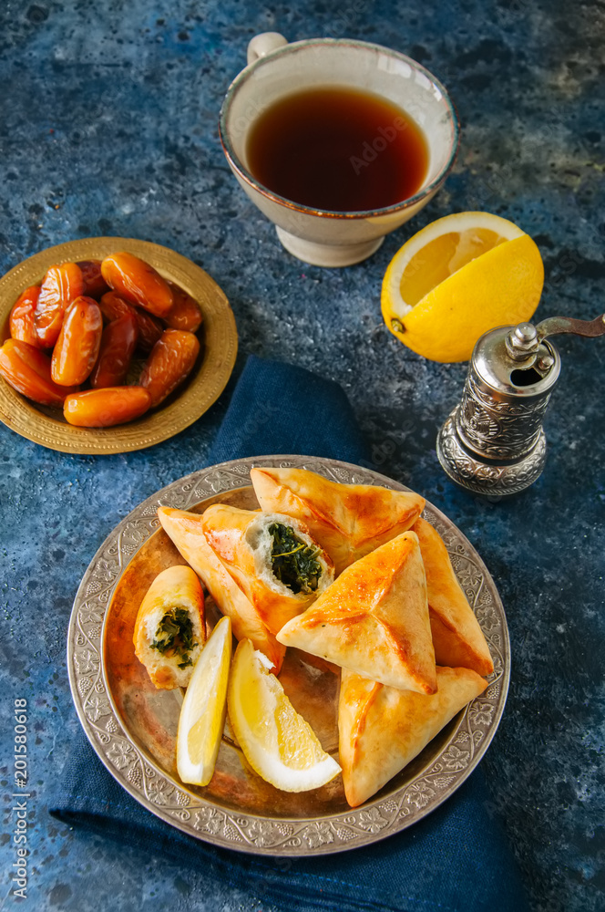 Iftar food during ramadan, arabic and middle eastern food concept. Fatayer sabanekh - traditional arabic spinach triangle hand pies dates and tea on a blue stone background. Top view. Iftar food during ramadan, arabic and middle eastern food concept. Fatayer sabanekh - traditional arabic spinach triangle hand pies dates and tea on a blue stone background. Top view.