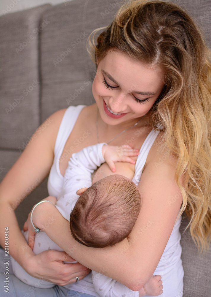 Breastfeeding baby. Pretty mother holding her newborn child. Mom smile and nursing infant. Beautiful woman and new born love at home. Blondе mother breast feeding baby.