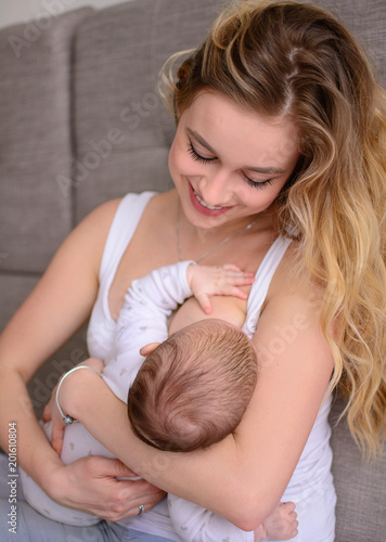 Breastfeeding baby. Pretty mother holding her newborn child. Mom smile and nursing infant. Beautiful woman and new born love at home. Blondе mother breast feeding baby.