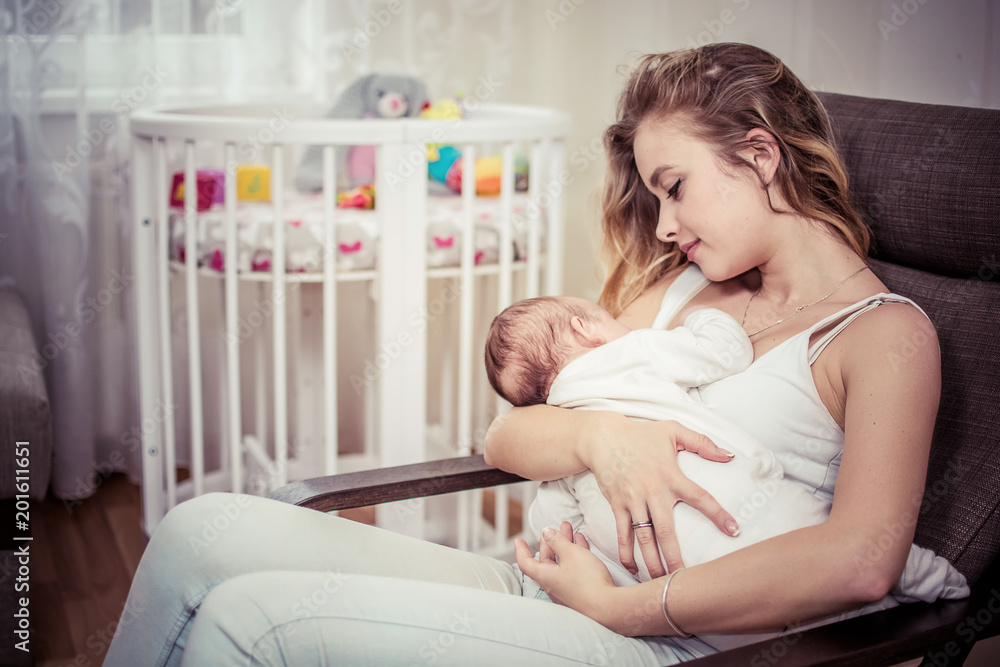 Breastfeeding baby near crib. Pretty mother holding her newborn child. Mom nursing baby. Beautiful woman and new born relax at home in nursery interior. Blondе mother breast feeding baby.