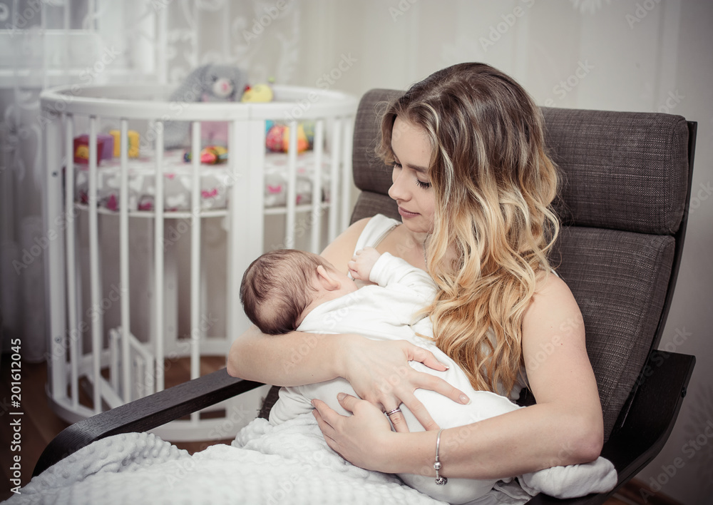 Breastfeeding baby near crib. Pretty mother holding her newborn child. Mom nursing baby. Beautiful woman and new born relax at home in nursery interior. Blondе mother breast feeding baby.