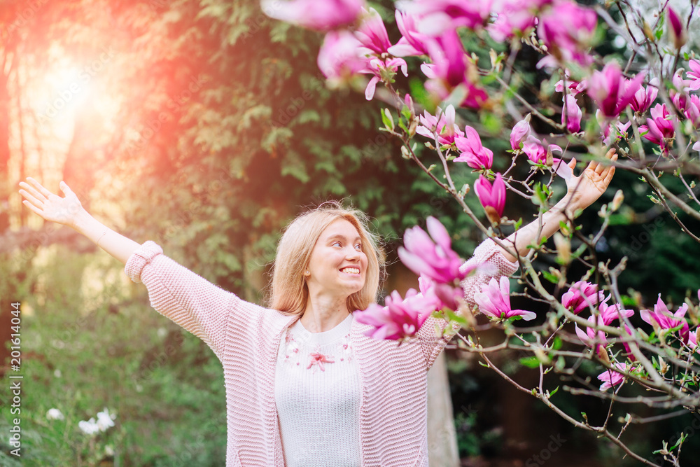 Excited european blond sweden woman raising her arms while standing near blossom magnolia tree in sunny spring morning. Sun glare effect.