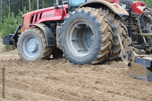 A tractor with a connected seeding unit, a combine, a drill with large wheels, travels across the field, sows crops, performs agrarian and farm work against the background of trees.