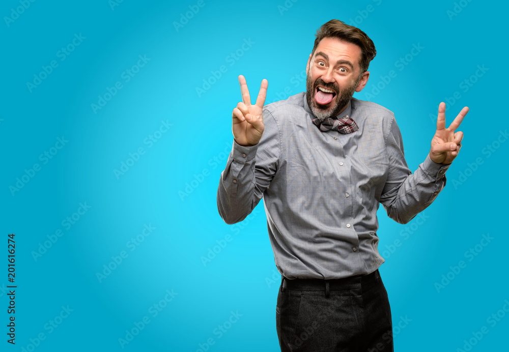 Fototapeta premium Middle age man, with beard and bow tie looking at camera showing tong and making victory sign with fingers