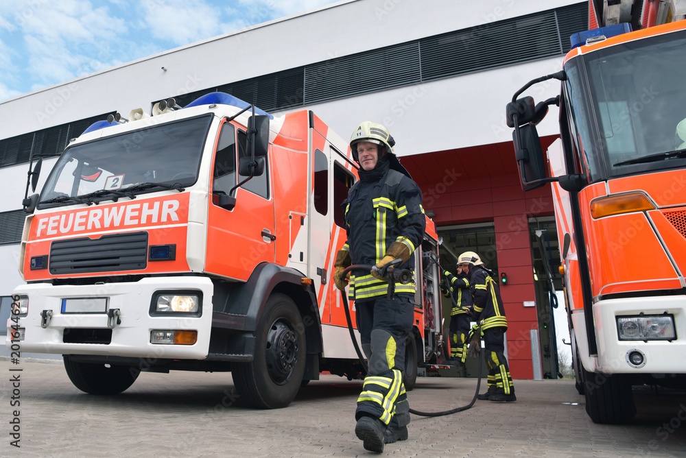 Fototapeta premium Feuwehrwehr: Feuerwehrmänner am Löschfahrzeug vor dem Einsatz zur Bekämpfung eines Brandes an der Feuerwache // Fire brigade: firefighters at the fire station