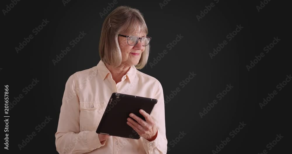 Charming senior woman using tablet device to browse online on gray background, Gentle mature lady using wireless pad device on dark grey backdrop, 4k Charming senior woman using tablet device to browse online on gray background, Gentle mature lady using wireless pad device on dark grey backdrop, 4k