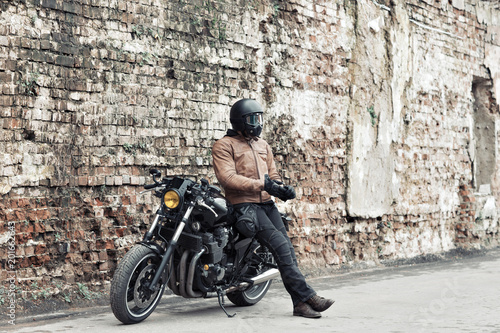 Man a motorcyclist standing with helmet near her bike, brick wall of garage background. Male in a leather jacket and tight pants photo