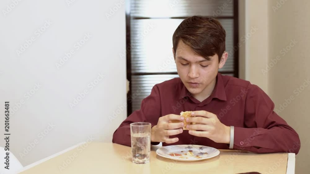 A 15 year old teen bites a sandwich. A young guy eats a sandwich at a table. A 15 year old teen bites a sandwich. A young guy eats a sandwich at a table.