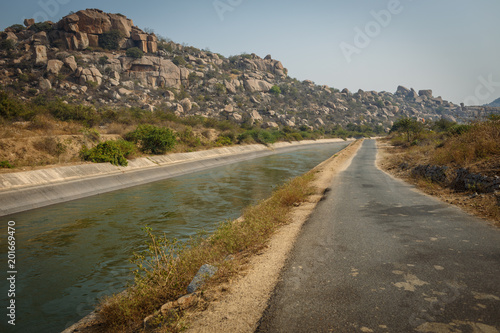 Asphalt road among mountains, boulders and rocks. A fast river flows along the road. Beautiful morning landscape. Uninhabited area. Hampi Village. India. Asia.