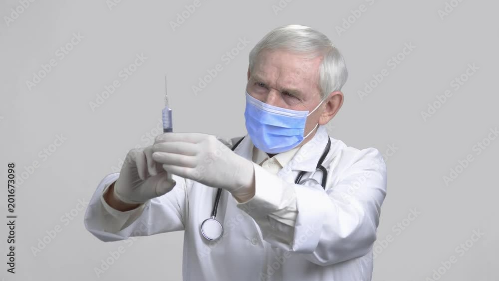 Old doctor with syringe in grey isolated background. Frown anesthetist wearing protective mask and stethoscope checking amount of water inside injector.