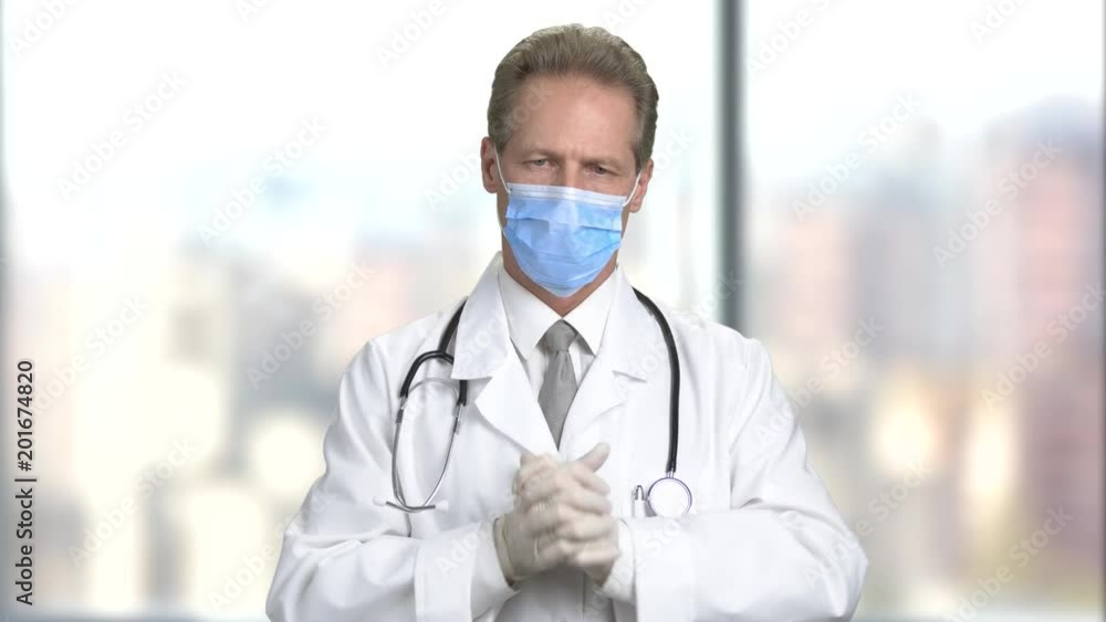 Serious doctor in mask put on gloves. Getting ready to surgery operation. Bright abstract blurred windows background with view on city. Serious doctor in mask put on gloves. Getting ready to surgery operation. Bright abstract blurred windows background with view on city.