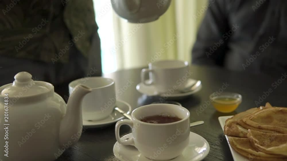 close up shot of the man's hands, the person pours a hot drink from the kettle into the porcelain cups, on the wooden table there are freshly baked pancakes and a container with sweet honey close up shot of the man's hands, the person pours a hot drink from the kettle into the porcelain cups, on the wooden table there are freshly baked pancakes and a container with sweet honey