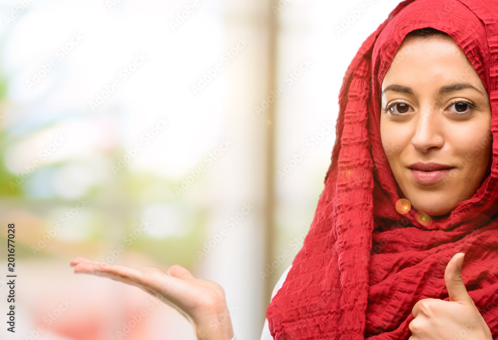 Young arab woman wearing hijab thumb up holding something in his empty hand and making ok gesture Young arab woman wearing hijab thumb up holding something in his empty hand and making ok gesture
