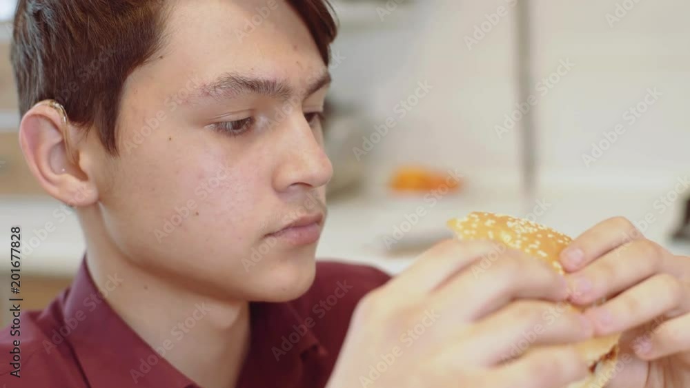 A 15 year old teen bites a sandwich. A young guy eats a sandwich at a table. A 15 year old teen bites a sandwich. A young guy eats a sandwich at a table.