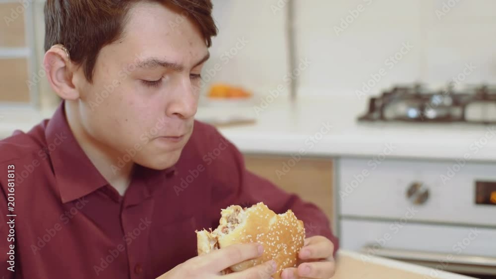 A 15 year old teen bites a sandwich. A young guy eats a sandwich at a table. A 15 year old teen bites a sandwich. A young guy eats a sandwich at a table.