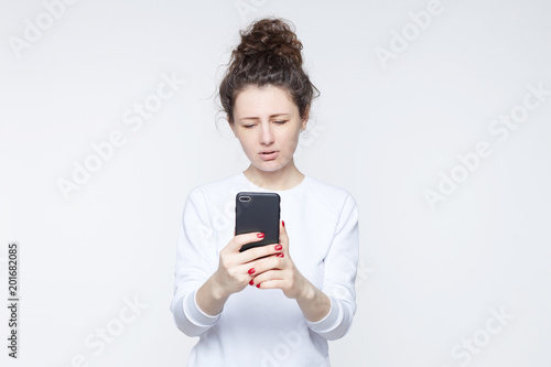 Close-up portrait of young American woman with curly hair in bun wearing casual T-shirt communicating over smartphone video, having sorrowful,sad expression. Human emotions and feelings concept.