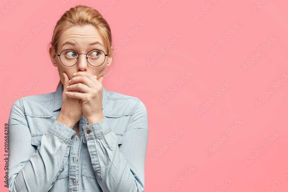 Scared female tries to be silent, keeps hands on mouth, looks with worried expression away, dressed in casual denim jacket, isolated over pink background with copy space for your advertisment
