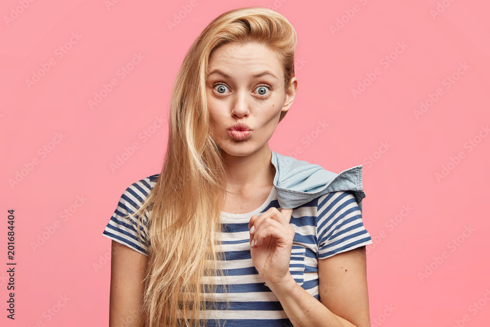 Portrait of attractive blonde young woman with pleasant appearance, wears striped t shirt, has round lips, stares at camera, makes grimace, holds jacket, isolated over pink studio background Portrait of attractive blonde young woman with pleasant appearance, wears striped t shirt, has round lips, stares at camera, makes grimace, holds jacket, isolated over pink studio background