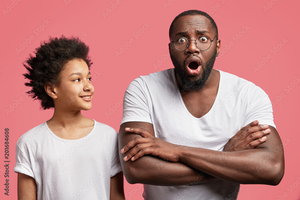 Happy teenage boy with Afro hairtyle looks with positive expression at his dark skinned surprised father who keeps mouth widely opened and arms folded, shocked to recieve bad unexpected news Happy teenage boy with Afro hairtyle looks with positive expression at his dark skinned surprised father who keeps mouth widely opened and arms folded, shocked to recieve bad unexpected news