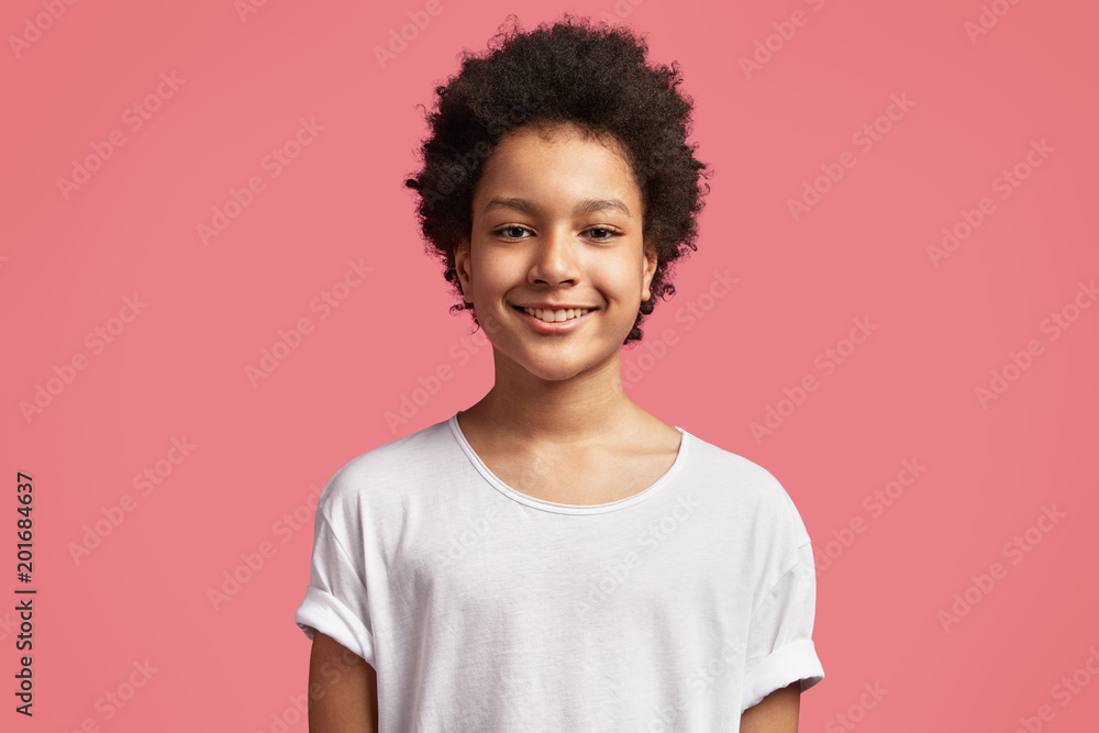 Waist up portrait of handsome smiling black mixed race teenager has Afro hairdo, dressed in casual white t shirt, being in good mood after playing with friends in yard outdoor during Saturday Waist up portrait of handsome smiling black mixed race teenager has Afro hairdo, dressed in casual white t shirt, being in good mood after playing with friends in yard outdoor during Saturday